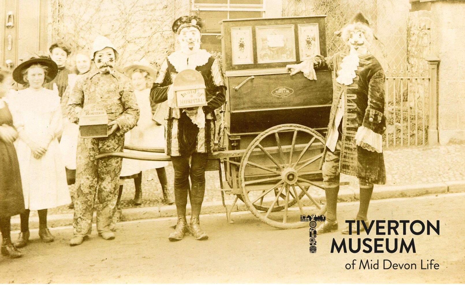 some people in costume standing round a barrel piano, an organ on wheels, in black and white