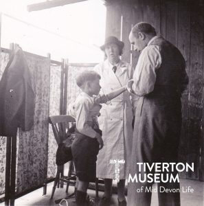 A black and white photo of a young boy holding his arm up. A man, presumably a doctor is holding the arm and looking at the boy. There is a woman in a long white coat and hat standing behind them. 