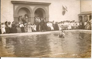 Two men in a swimming pool. One is dragging the other backwards through the water demonstrating an old lifesaving technique to onlookers. 