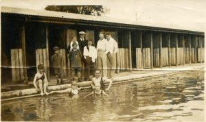 A group of people in a swimming pool looking at the camera with several more standing behind them on the poolside in front of a row of changing cubicles