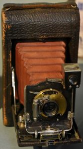 An old camera: a brown leather box with a tunnel of red fabric coming out of the front connected to a metal disc where the lens sits. 