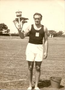 A man wearing a dark sleeveless top and white shorts standing in a playing field holding a metal torch