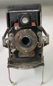 The front of an old camera. A black box propped up on two metal stands, with a hole in the middle where the lens sits, surrounded by a rusty metal disc