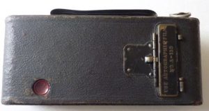 An old camera in the shape of a dark brown, leather, rectangular box with a small red button on the bottom left, a handle at the top and a metal clasp on the right