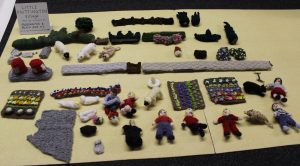 A group of knitted people and animals laid out on a table. There are also some knitted vegetable patches and other accessories. There is a handwritten sign saying 'Little Knittington Village'.