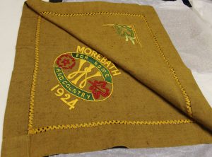 A piece of dark green or brown cloth. 'Morebath for home and country 1924' is embroidered in yellow thread as part of an emblem featuring some red flowers. There is a leaf motif embroidered in yellow and green in the opposite corner.