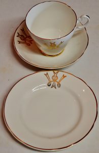 A white or cream cup and saucer and a side plate, all edged in gold. They all bear an emblem with a letter W and a letter I intertwined.