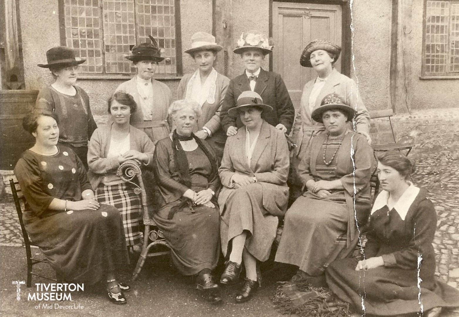 A black and white photo showing 6 seated women with 5 women standing behind them, in front of a building. The women are dressed in Edwardian fashion with loose, flowing clothing (dresses and skirts). Several of the women are wearing hats. The women look relaxed and most are smiling.