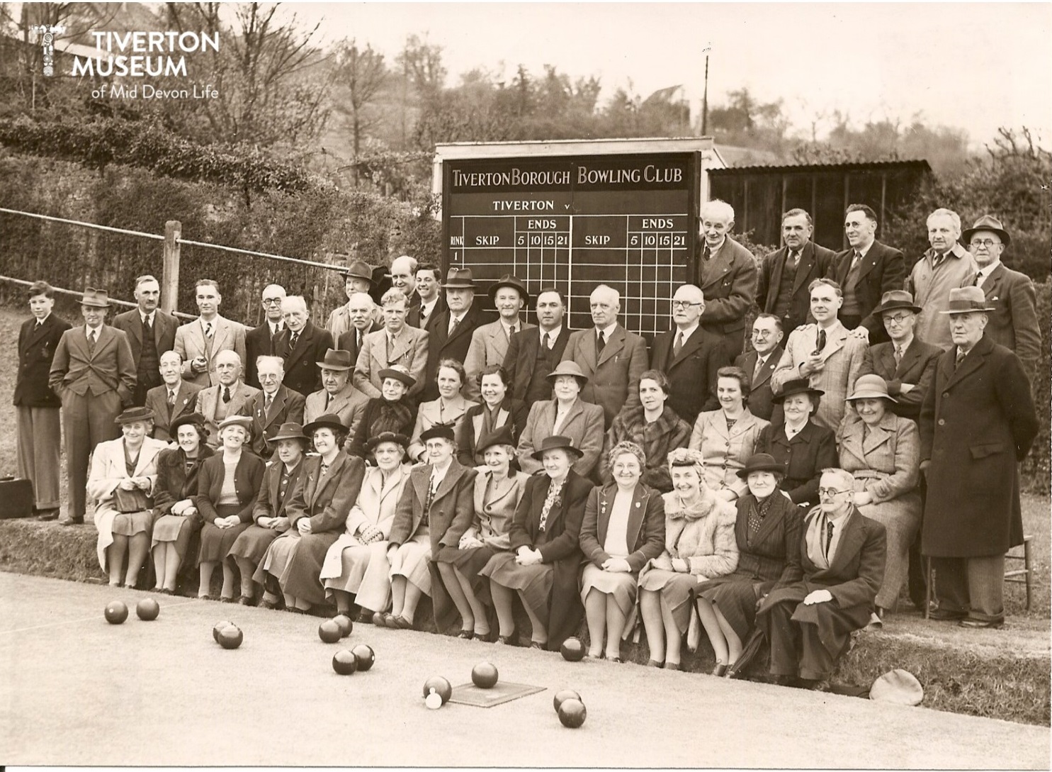 A group of men and women seated and standing in three rows on the edged of a bowling green in front of a board that reads 'Tiverton Borough Bowling Club' with a system for keep score below. The woman are all wearing overcoats and skirts and the men are wearing overcoats or jackets. Lots of the women and some of the men are wearing hats. There are some bowling balls on the ground in front of them.
