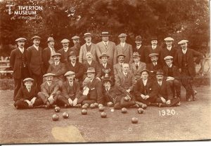 An old black and white photo of a group of men in jackets and hats gathered for a photo in front of some trees on a bowling green. They are arranged in three rows, standing at the back, seated in the middle and sitting in the front. There are bowling balls in front of them and the man seated in the middle is holding a cup. '1920' has been written on the bottom right of the photo. Lots of the men have large moustaches.