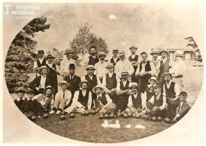 An old black and white photo of a group of men in front of a pavilion and some trees in a park. Some of the men are kneeling or seated in front with a small number of young boys also kneeling. There are bowling balls at their feet. The men are dressed in late Victorian fashion with white shirts and either dark waistcoats or jackets. All the men are wearing hats, several have large moustaches and a few are smoking pipes.
