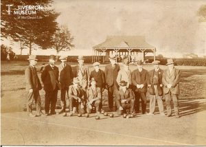 An old black and white photo showing a group of men in front of a pavilion in a park. The men are all wearing jackets and hats. Three of the men are kneeling in the front with a number of bowling balls in front of them.