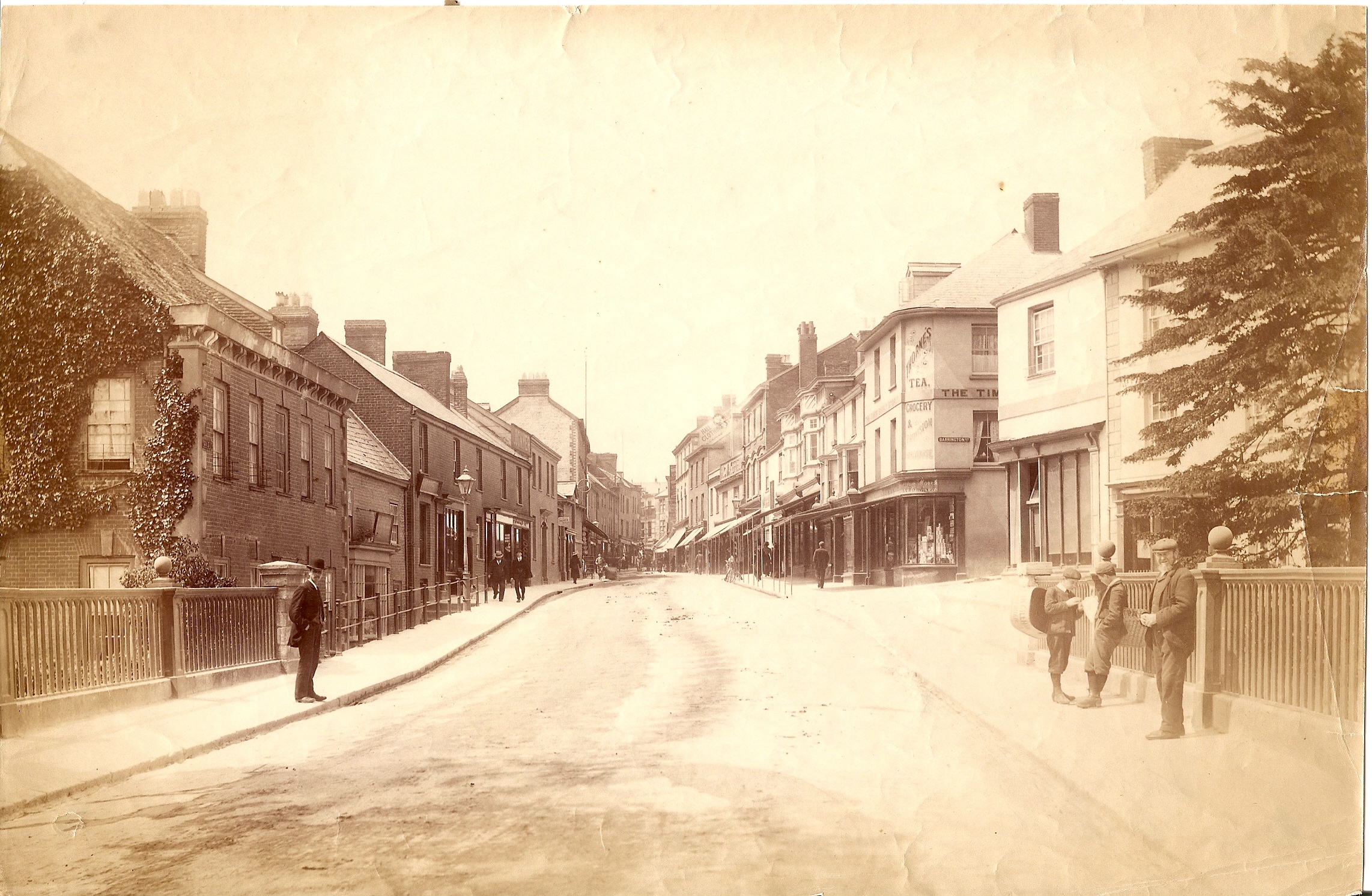 An old black and white photo with a sepia tint. It shows a view up Tiverton's Gold Street in around 1900, with a wide road and buildings either side. The building on the left has ivy climbing up the side and there is a big tree on the right of the image. There are several men standing around on either side of the street in Victorian dress; smart jackets and hats.
