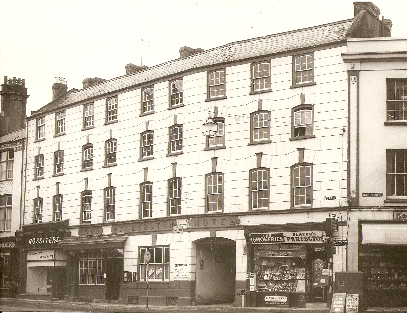 A black and white photo showing a large hotel building, Lord Palmerston Hotel. It is a white four storey building with lots of black framed windows and an entrance for an old fashioned coach and horses in the middle of the ground floor. There are also a couple of shop fronts on the ground floor to either side of the coach opening, Rossiters Opticians on the left and a tobacconist on the right.