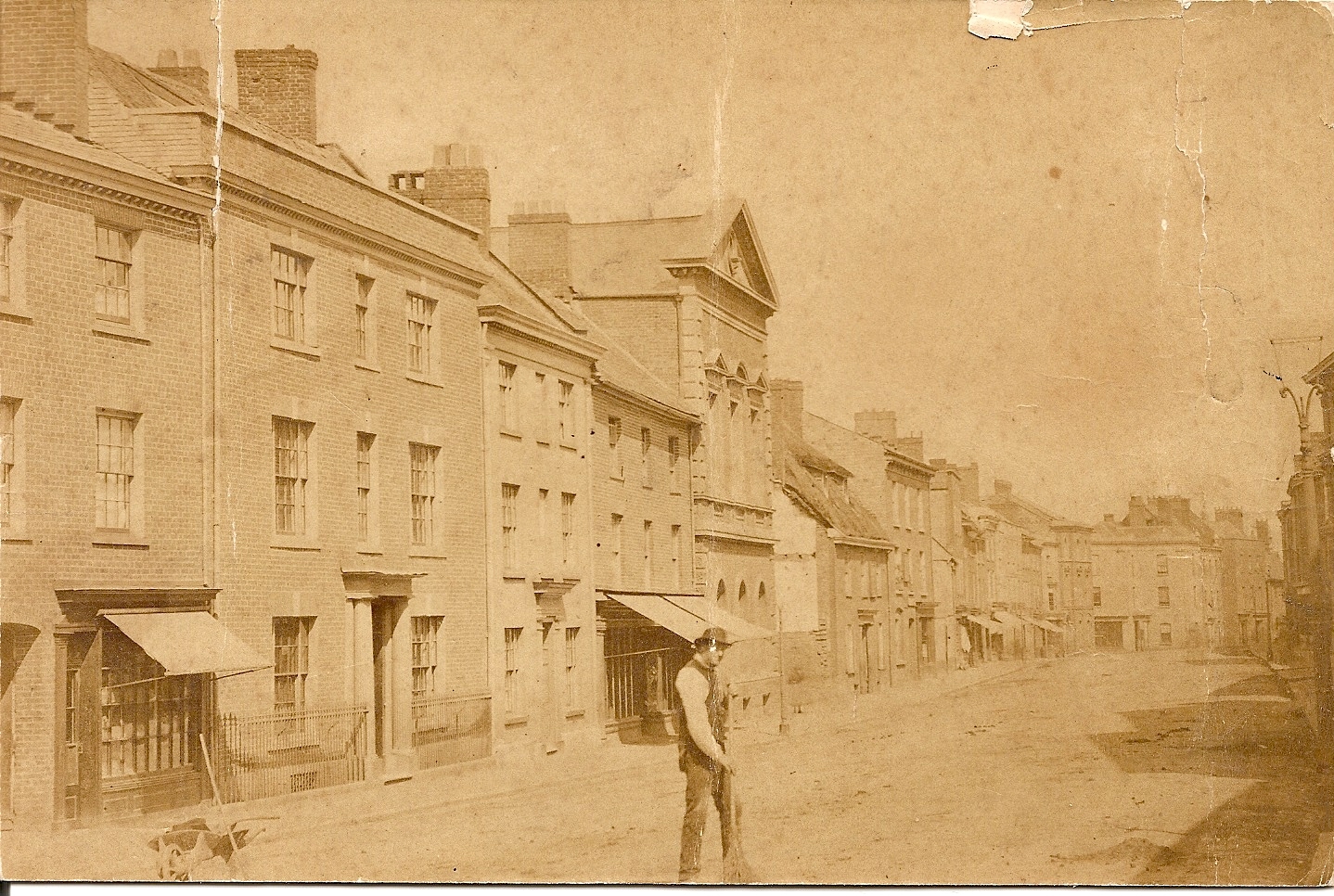 An old black and white photo a brown tint and other signs of age like a tear in one corner. It shows an unpaved road with three storey townhouses. and awnings over shopfronts. There is a wheelbarrow on the left of the image with some tools in and a man standing in the middle of the street holding what looks like a rake. He is wearing a white shirt with a waistcoat over the top and a bowler hat. The photo is recorded as showing Crediton high street around 1900.