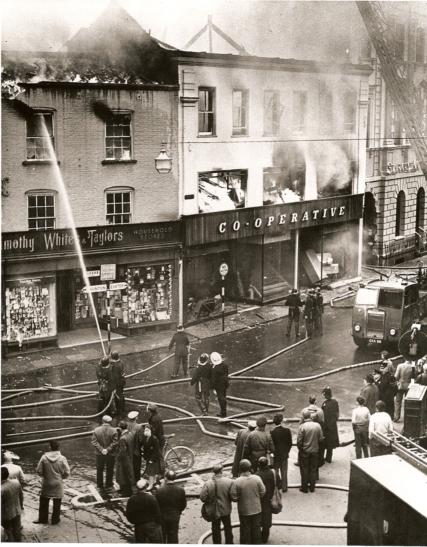 A black and white photo dated from around 1960. It shows two buildings on Tiverton's Fore Street, Whites & Taylors Household Stroes and Co-Operative, on fire. There are firefighters in the foreground tackling the blaze with hoses and lots of bystanders watching the fire.
