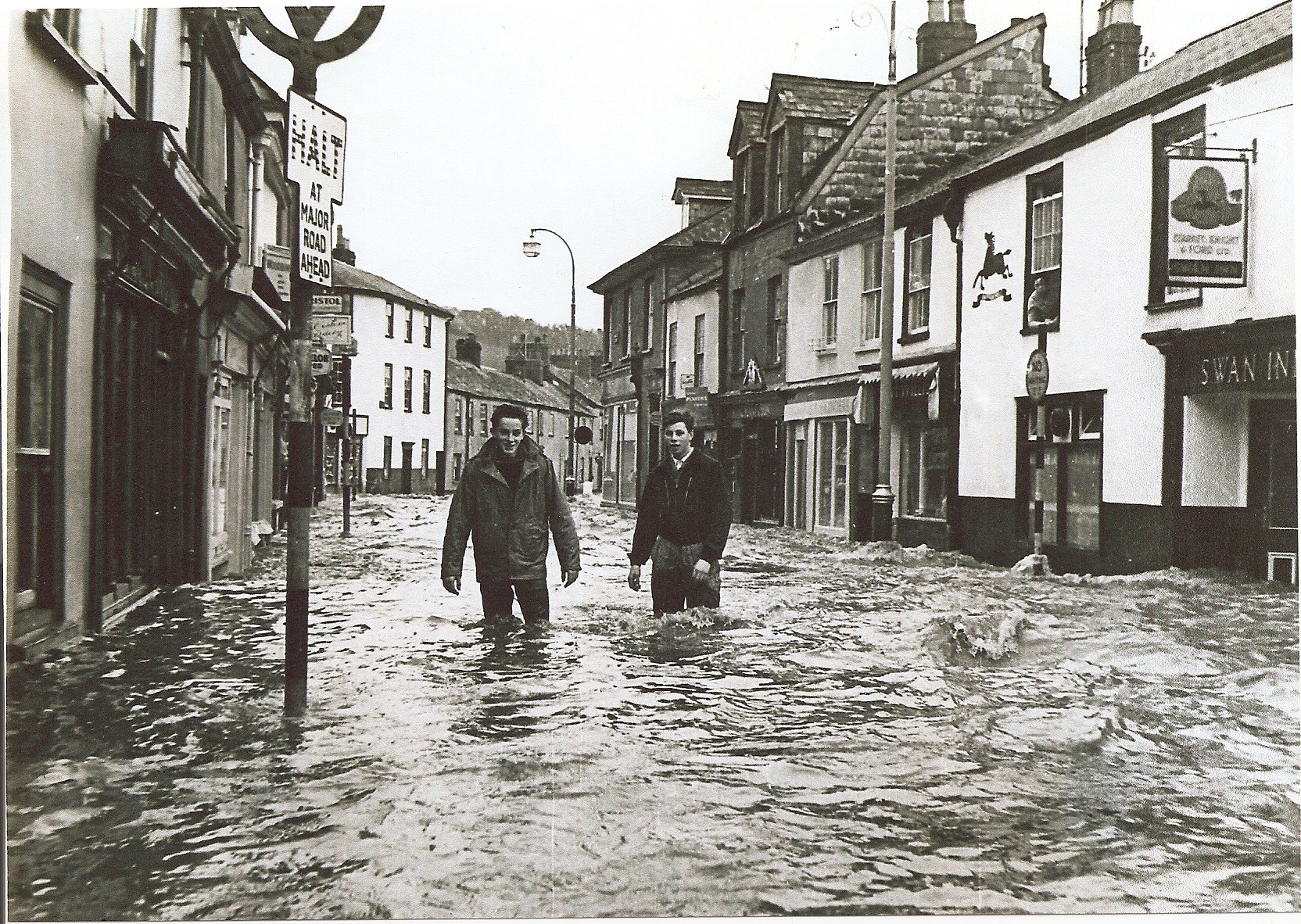 A black and white photo dating from 1960. A flooded street with a pub, the Swan Inn, on the right and building either side. There are two men standing in the middle of the street. The flood water reaches just over their knees.