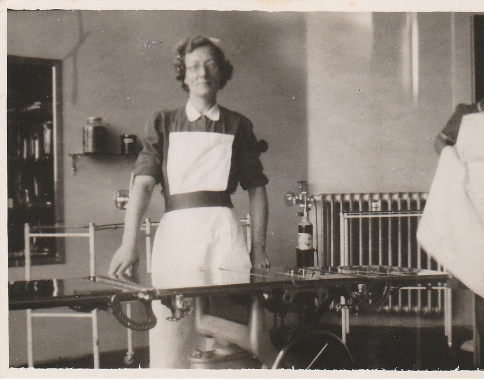 A black and white photo showing a female nurse in uniform standing behind a medical table.