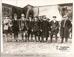 An old black and white photo showing a group of men, wearing old fashioned riding breeches and smart jackets. The mean are all wearing hats, some flat caps, some trilbys. Some of the men have smoking pipes in the mouths. They are standing in front of a wall that has ivy climbing up it. 