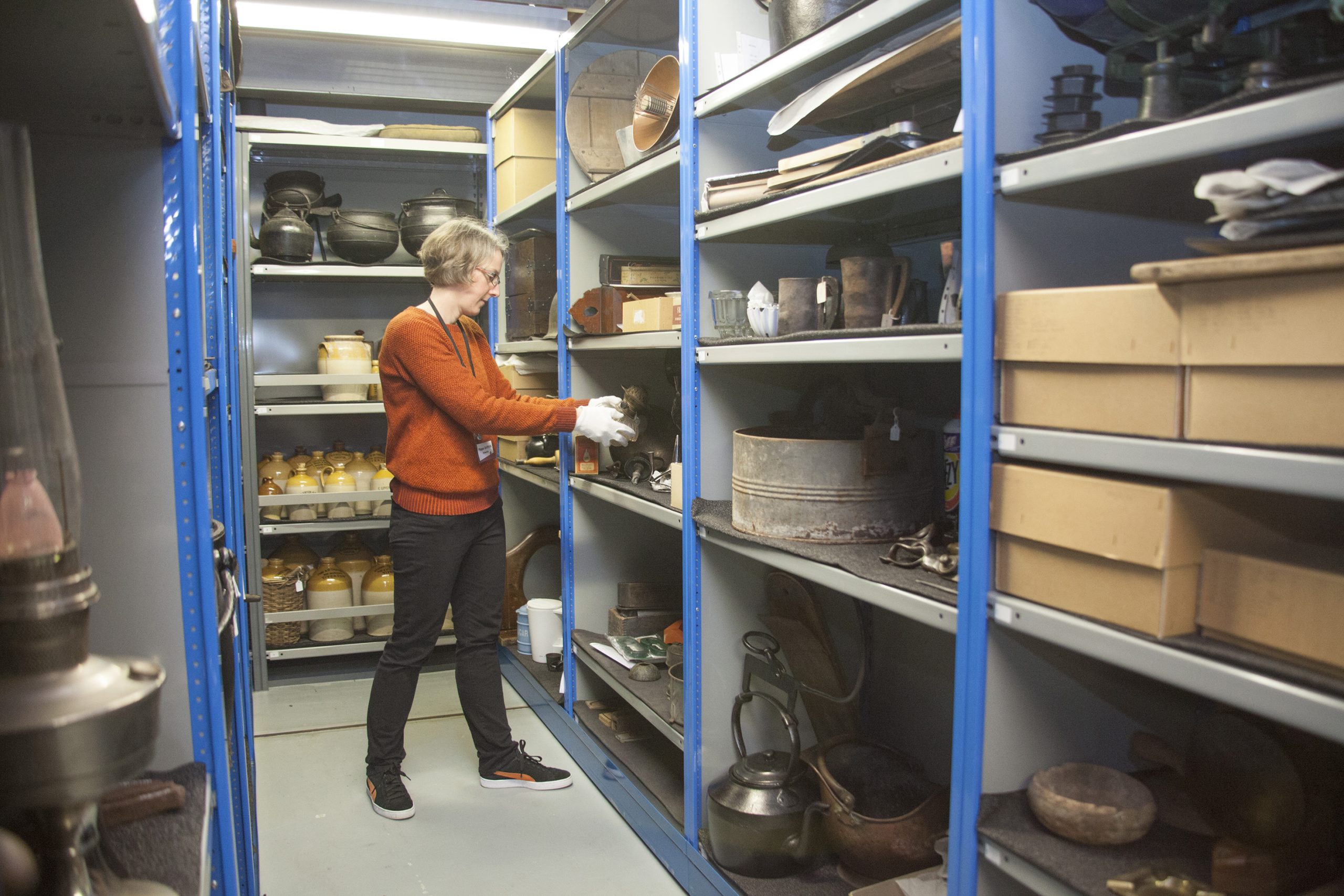 A woman in an orange jumper and black trousers standing in a museum store. She is wearing white cotton gloves and placing a museum object on a rack of shelving which has other museum objects and boxes on.