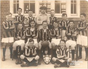 An old black and white group photo of a boy's football team. There are 3 rows of boys, 7 boys standing, 5 boys seated in the middle and two sitting cross legged in the front row. The boys are dressed in white shorts and striped football jerseys. One boy at the back of the group is wearing a light coloured jumper and a flat cap. There is a shield shaped trophy on the floor between the two seated boys.