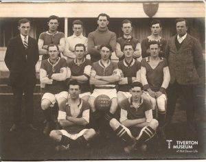 An old black and white group photo of a football team. There are 3 rows, 7 men standing at the back, 5 men seated in the middle and two men sitting cross legged on the floor in the front. Most of the men are wearing shorts and t-shirts with football boots and long socks. One man is wearing a woollen jumper and two are wearing suits. There is a ball between the legs of the man seated in the middle of the group. There is writing on the ball that reads Tiverton AFC 1922 - 23.