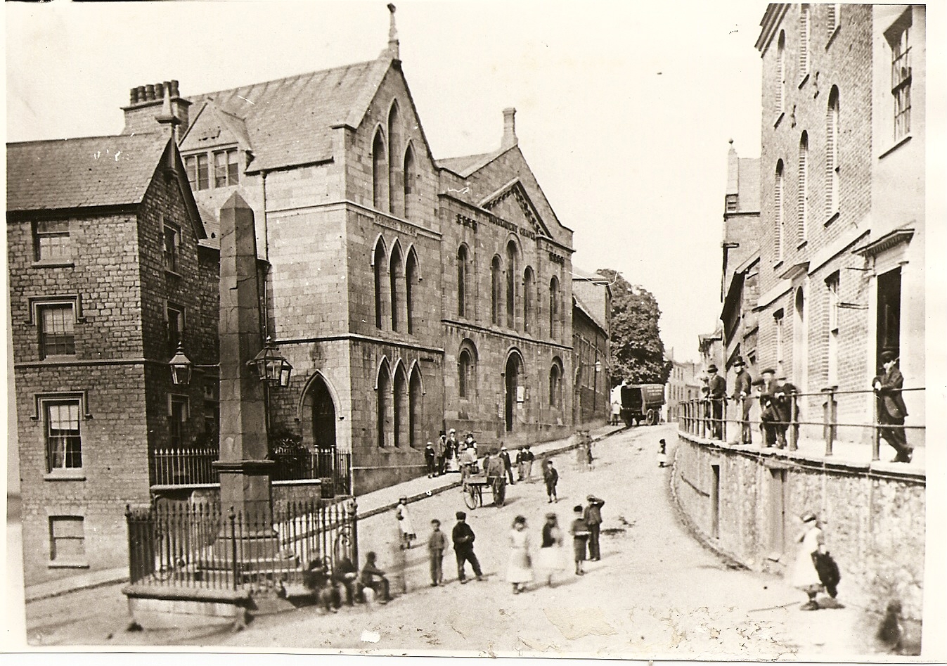 A street scene in black and white with large bulidings in the background and an obelisk in the foreground. Ther are several people milling around. The photo is dated 1840.