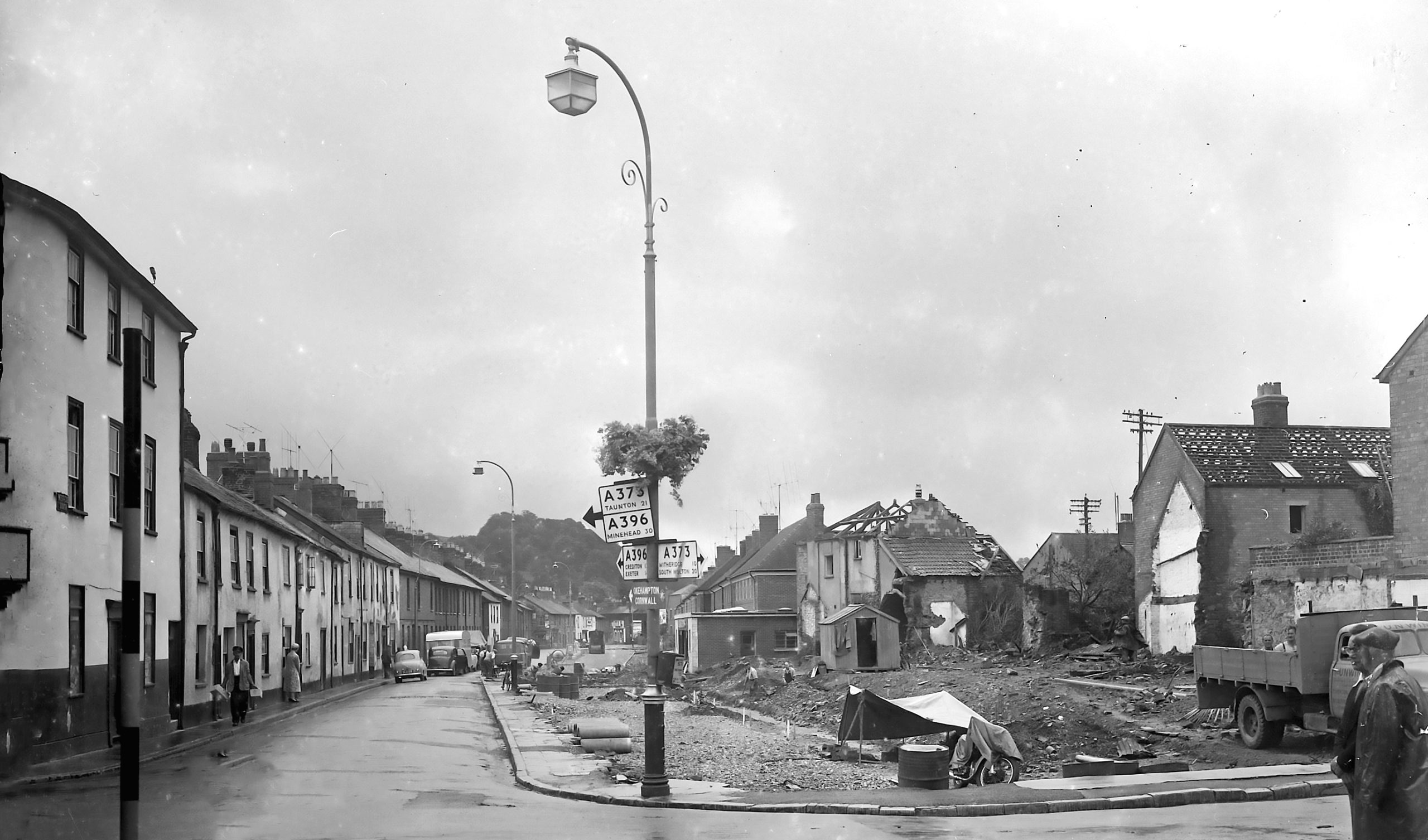 Black and white photo showing a street scene with some demolished houses next to a road.