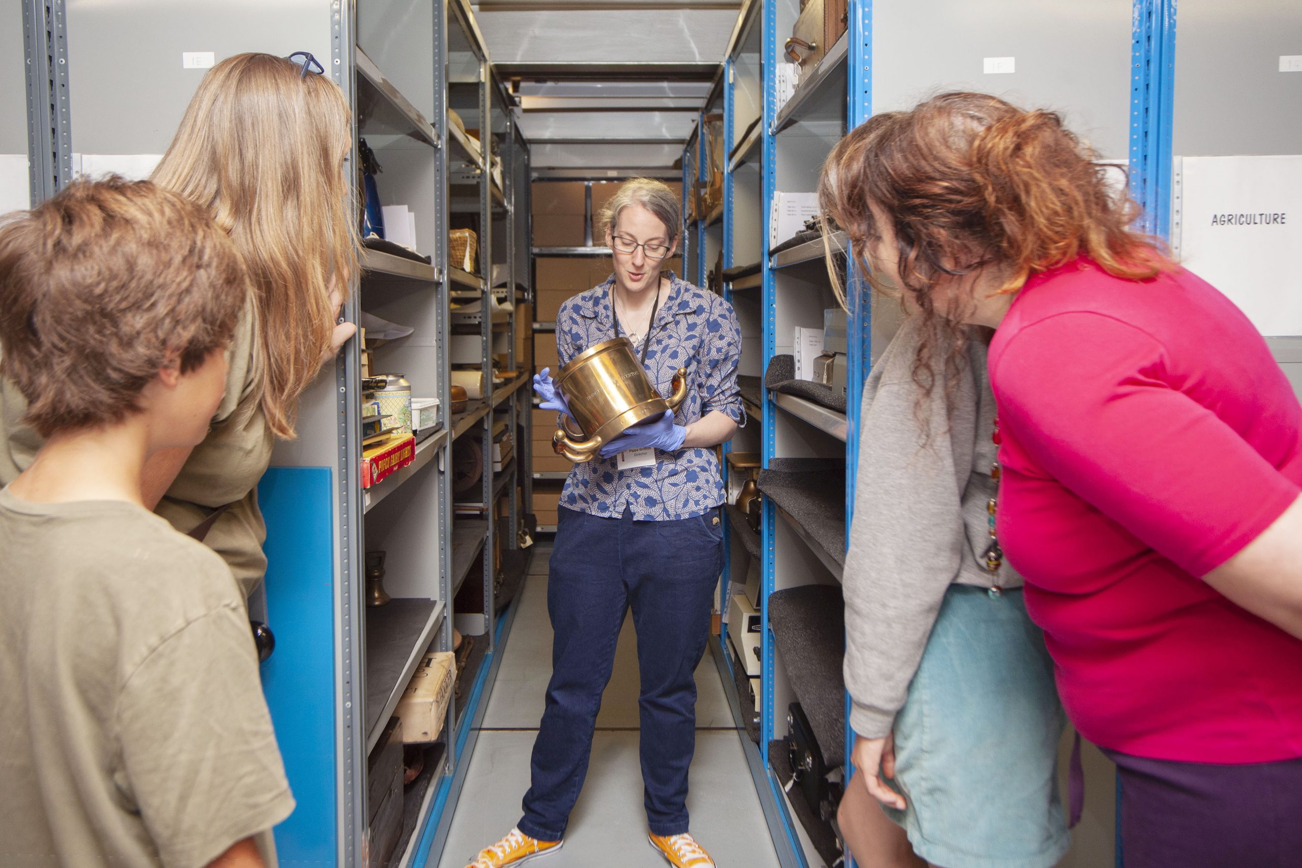 A woman standing in between some shelving units in a museum store, facing the camera. She is holding a brass object and there are other museum objects on the shelves around her. There are people peering around the end of the shelves looking at the woman.