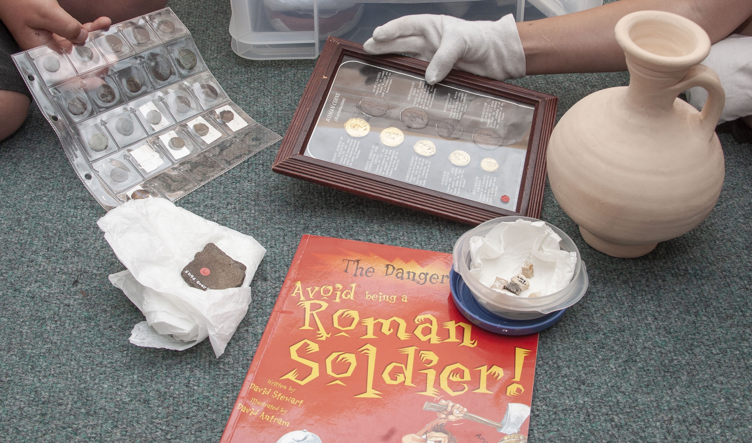 Hands wearing white gloves holding a frame with replica coins in. There are other museum objects on the floor around the hands, including a ceramic jug.