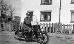 A black and white photo of a man sitting on a motorbike in front of a house. The bike has an AA logo, as does the man's helmet. He is wearing a big heavy looking coat and gloves.