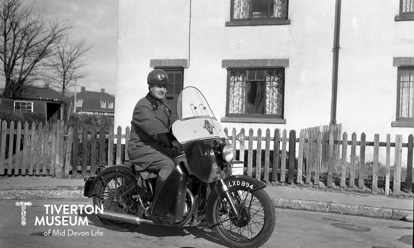 A black and white photo of a man sitting on a motorbike in front of a house. The bike has an AA logo, as does the man's helmet. He is wearing a big heavy looking coat and gloves.