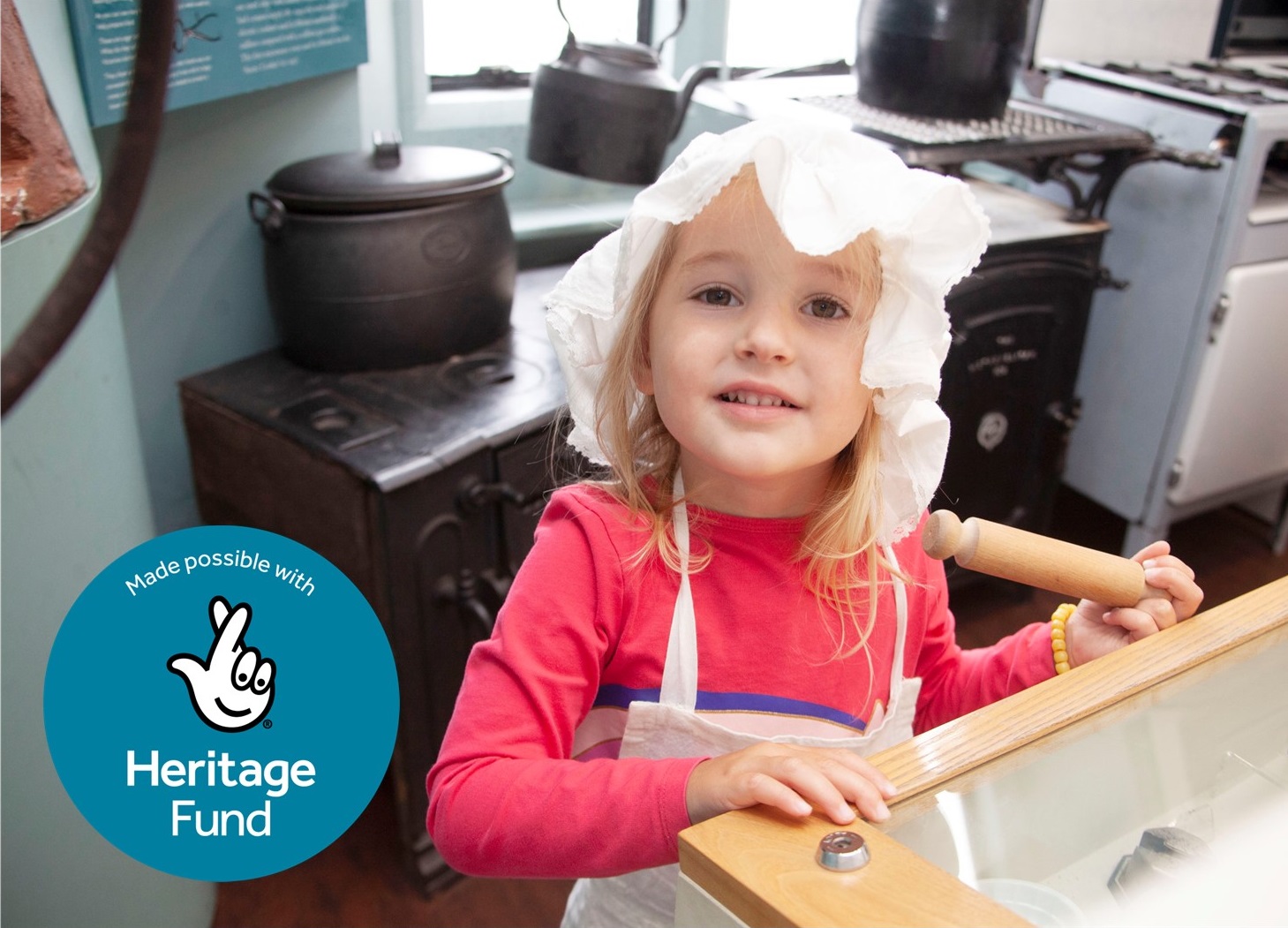 A young white girl with blond hair is looking at the camera smiling. She was wearing a dressing up apron and mob cap and holding a toy rolling pin. There is a blue circle overlaid on the left hand side of the photo with a hand with crossed fingers (the National Lottery logo) ad the wording 'Made possible with Heritage Fund'.