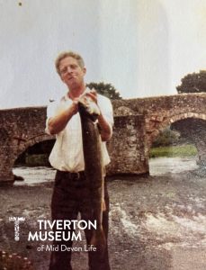 A man standing on a river bank holding a large pike fish. He is wearing a white short sleeved shirt and brown trousers and has a cigarette hanging out of his mouth. There is an old stone bridge behind him. The fish must be heavy because he is holding it with two hands.