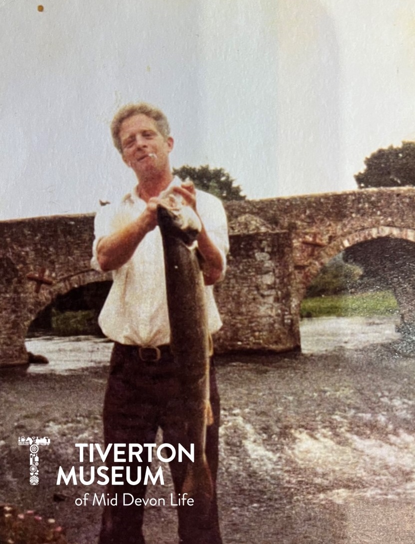 A man standing on a river bank holding a large pike fish. He is wearing a white short sleeved shirt and brown trousers and has a cigarette hanging out of his mouth. There is an old stone bridge behind him. The fish must be heavy because he is holding it with two hands.