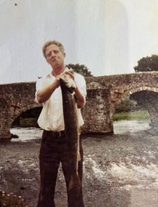 A man standing on a river bank holding a large pike fish. He is wearing a white short sleeved shirt and brown trousers and has a cigarette hanging out of his mouth. There is an old stone bridge behind him. The fish must be heavy because he is holding it with two hands.