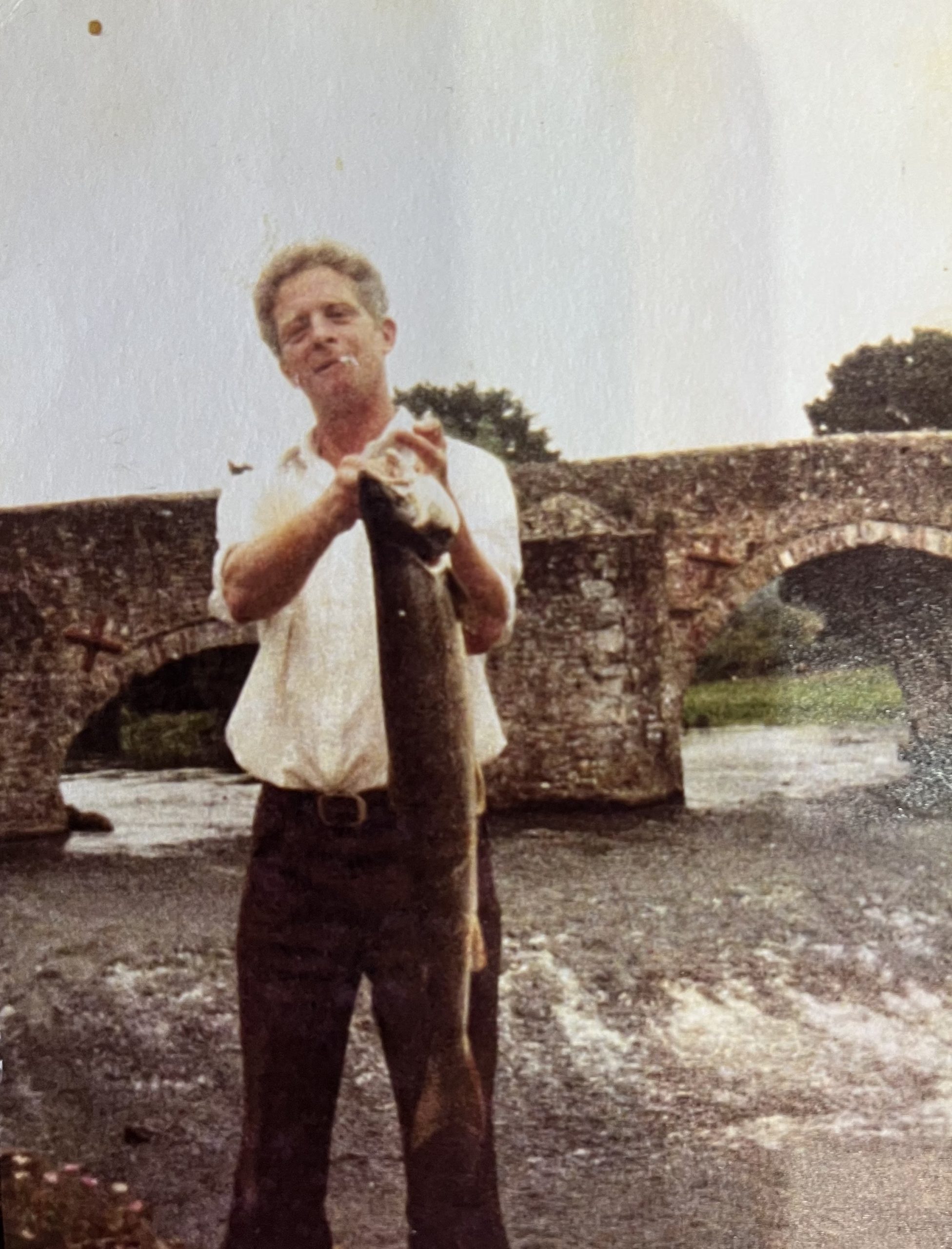 A man standing on a river bank holding a large pike fish. He is wearing a white short sleeved shirt and brown trousers and has a cigarette hanging out of his mouth. There is an old stone bridge behind him. The fish must be heavy because he is holding it with two hands.