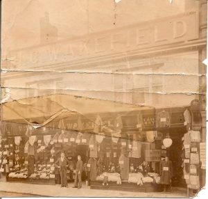 An old and creased photo showing a shopfront with a large sign reading 'H. C. Wakefield'. There are twon men standing in the doorway. There are lots of pieces of clothing hanging up on display outside the shop.