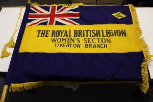 A blue banner with gold coloured fringe, laid out on a table. There is a union jack flag in the top left hand corner and a gold strip across the middle with the words 'The ROyal Britisih Legion Women's Section Tiverton Branch' embroidered in blue. In the top right hand corner there is a gold coloured diamond shape with '85 years' embordered in blue.