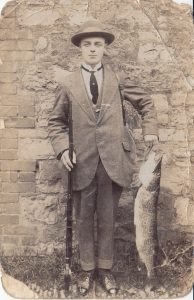 A man standing in front of a brick wall holding a fish on a hook. He is wearing a suit wity jacket and tie, shiny shoes and a hat. The photo is black and white and is dated 1879, the clothes are of this period. He is holding the fish at about waist height and it reaches all the way to the floor.