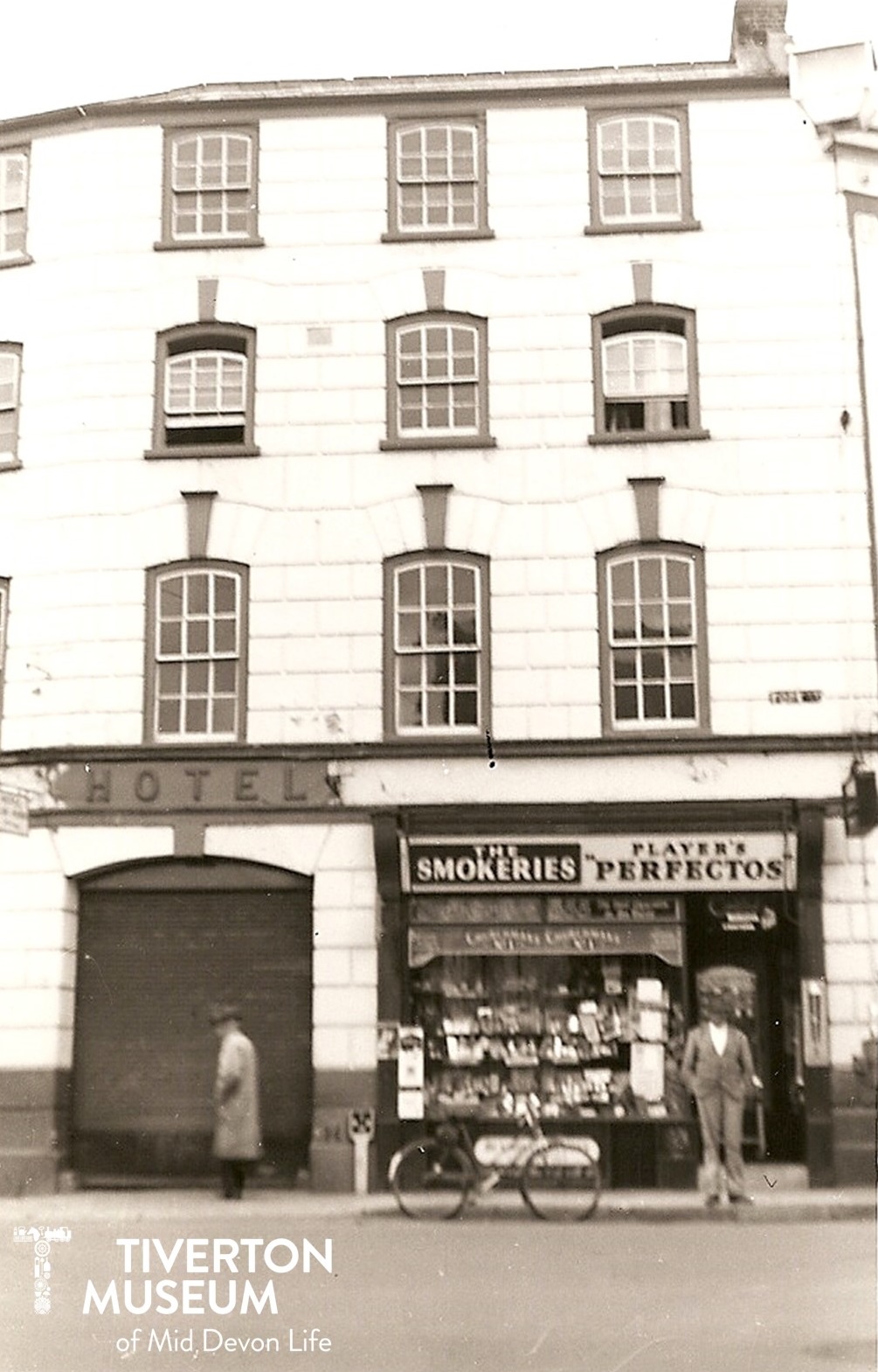 Black and white image of a four storey building with large windows. On the bottom left there is a sign saying 'HOTEL' with a large arch underneath. There is a man in a long overcoat walking past the arch. On the bottom right is the front of a shop with a man standing outside looking at the camera. The shop sign reads 'The Smokeries' and the window display is very full but it's too far away to see what the products are. There is a bicycle propped up against the kerb in front of the shop and the man is wearing a light coloured suit.