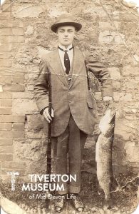 A man standing in front of a brick wall holding a fish on a hook. He is wearing a suit wity jacket and tie, shiny shoes and a hat. The photo is black and white and is dated 1879, the clothes are of this period. He is holding the fish at about waist height and it reaches all the way to the floor.