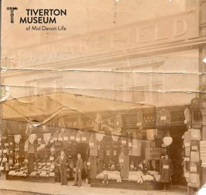 An old and creased photo showing a shopfront with a large sign reading 'H. C. Wakefield'. There are twon men standing in the doorway. There are lots of pieces of clothing hanging up on display outside the shop. 