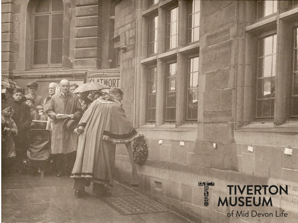 A man in a long gown laying a wreath outside a building. He is facing away from the camera and you can see part of a crowd gathered on the other side of him, facing towards the camera.