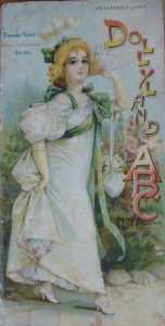 A card featuring an illustration of a young woman with blonde curly hair wearing a long white dress which she is lifting slightly to reveal her white, heeled shoes. The text reads 'Dollyland ABC', 'Father Tuck's ever welcome series' and 'untearable linen'. 