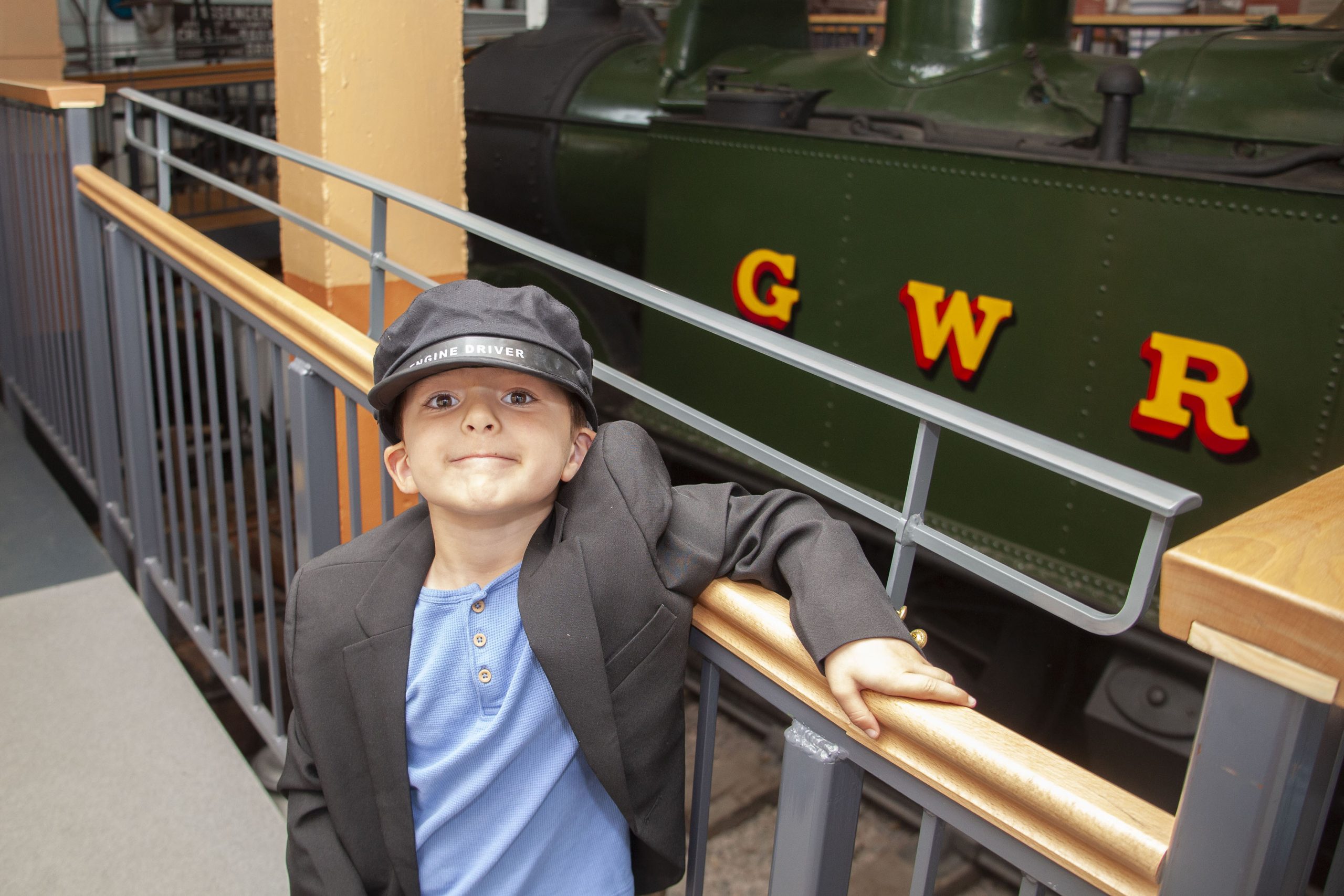 A young boy wearing a dressing up black jacket and an engine driver's black cap, standing in front of a green steam engine inside a museum gallery.