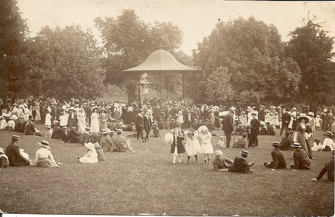 A park with a bandstand in the background. There are crowds of people sitting, standing and walking in the park