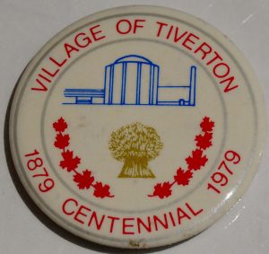 A commemorative badge pin, white background with red writing in a circle round the edge reading 'Village of Tiverton - 1879 Centennial 1979'. In the centre there is a wheatsheaf, with a large building above and some maple leaves underneath. 