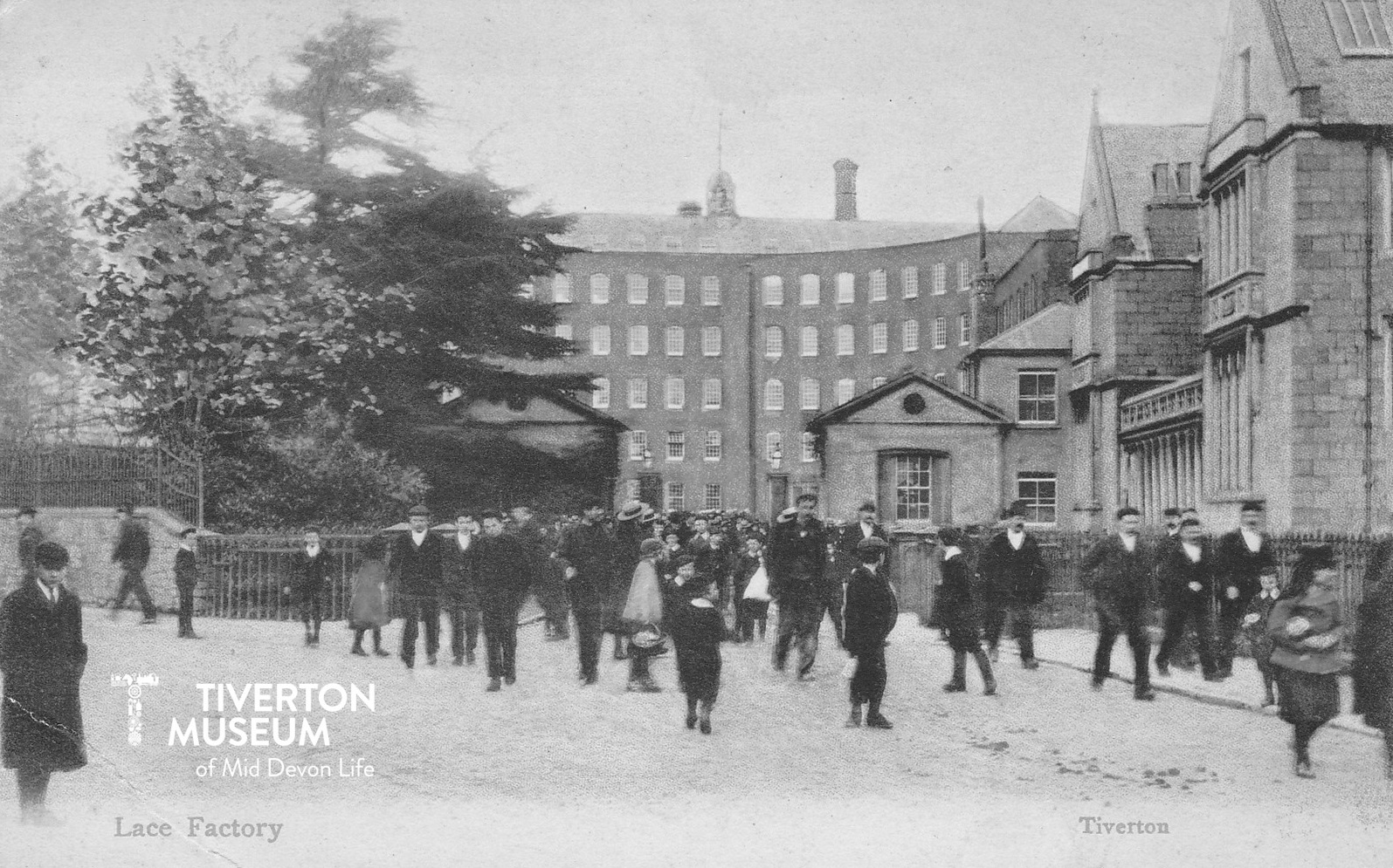 A large crowd of people leaving a large factory building with lots of small windows. The crowd is mostly men and boys and they are in old fashioned dress, mostly dark jackets and hats. The photo is in black and white.