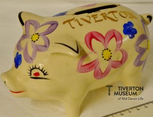 A ceramic 'piggy bank'. It is cream coloured with some bright flowers painted on the sides and 'Tiverton' painted near the slot for coins. 
